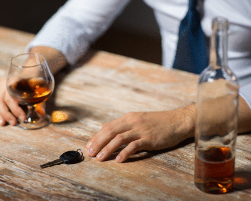 A mans hands on top of a wooden bar holding a car key in one hand and a glass of whiskey in the other.