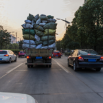 Overloaded cargo truck carrying stacked bags drives along a roadway with surrounding traffic, illustrating the risks of overweight commercial vehicles on public roads.