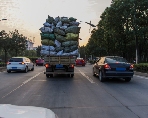 Overloaded cargo truck carrying stacked bags drives along a roadway with surrounding traffic, illustrating the risks of overweight commercial vehicles on public roads.