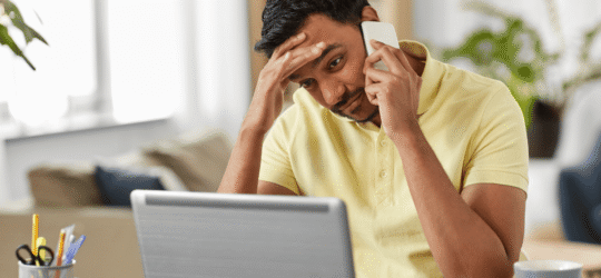 Stressed person on a phone call with a hand on their forehead while looking at a laptop, illustrating the difficulty of dealing with insurance agent contact after accident Colorado Springs.