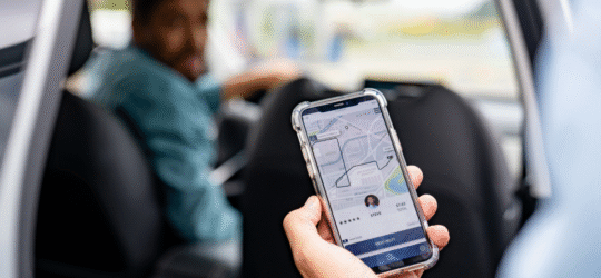 A passenger holds a smartphone displaying a rideshare app while sitting in the back seat of a car, with the driver visible ahead, illustrating rideshare transportation and the legal differences between Uber or Lyft accidents and private car crashes in Colorado Springs.