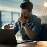Man sitting at a desk with a laptop, holding his glasses and rubbing his eyes in frustration, symbolizing the stress and concern over whether restraining orders appear on Colorado background checks and how they affect employment or housing opportunities.