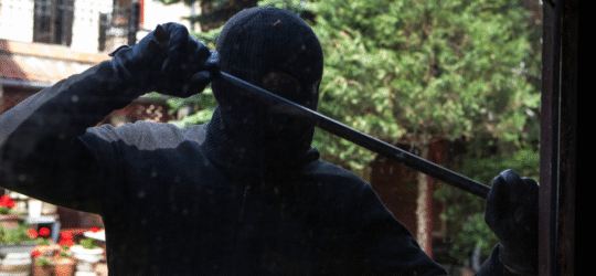 Man dressed in all black including gloves and a ski mask uses a crow bar to pry something open.