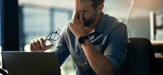 Man sitting at a desk with a laptop, holding his glasses and rubbing his eyes in frustration, symbolizing the stress and concern over whether restraining orders appear on Colorado background checks and how they affect employment or housing opportunities.