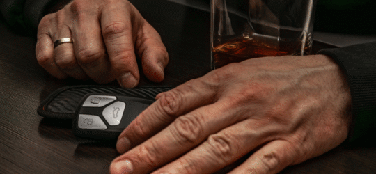 Close-up of a person’s hands resting on a table beside a glass of liquor and car keys, representing the professional and legal consequences of a DUI conviction in Colorado, including mandatory license reporting and disciplinary action by state licensing boards.