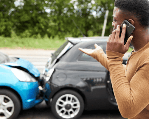 Frustrated driver on phone after rear-end collision between two compact cars, with visible damage to both vehicles, highlighting the aftermath of a traffic accident and the need for legal or insurance assistance