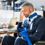 Injured man in neck brace and wheelchair holding crutches, sitting in an office with a concerned expression while meeting with a legal or medical professional, illustrating the aftermath of a personal injury and potential claim consultation