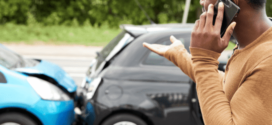 Frustrated driver on phone after rear-end collision between two compact cars, with visible damage to both vehicles, highlighting the aftermath of a traffic accident and the need for legal or insurance assistance